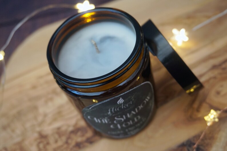 May include: A brown glass jar candle with a black label that reads "Flicker & Co. The Shadow Cloud Soy Wax Candle". The candle is lit and the wax is melting.