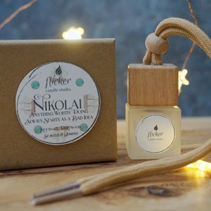 Puede incluir: Una caja de cartón marrón con una etiqueta blanca que dice "flicker candle studio. NIKOLAI ANYTHING WORTH DOING ALWAYS STARTS AS A BAD IDEA ROCKROSE,  DRIFTWOOD,  SCENTED & LINEN" y una pequeña botella de madera con una tapa marrón y una etiqueta blanca que dice "flicker candle studio." La botella está atada a una cuerda marrón.