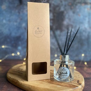 Könnte beinhalten: Eine klare Glas-Diffusorflasche mit schwarzen Schilfrohren und einem silbernen Deckel steht auf einem Holzbrett. Die Flasche hat ein Etikett mit der Aufschrift "Jack" und "Hot Chocolate". Daneben steht eine hohe, braune Pappschachtel mit dem "flicker"-Logo.