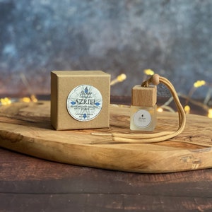 May include: A small brown box with a white label that reads "Flicker Azriel", a natural cedar and musk fragrance. The box is next to a small glass bottle with a wooden top and a beige string. The bottle is filled with a clear liquid and has a white label that reads "Flicker".