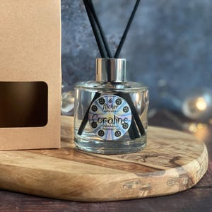 Pu&ograve; includere: Un diffusore in vetro trasparente con bastoncini neri e un tappo argentato, etichettato "Coraline" con un disegno floreale. La bottiglia &egrave; appoggiata su una tavola di legno accanto a una scatola di cartone marrone con una finestra.