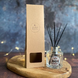Könnte beinhalten: Ein klarer Glasdiffusor mit schwarzen Stäbchen und einem silbernen Deckel, der mit "Maya" beschriftet ist. Er steht auf einem Holzbrett neben einer hohen, braunen Pappschachtel mit einem Logo. Der Hintergrund ist verschwommen blau und braun.