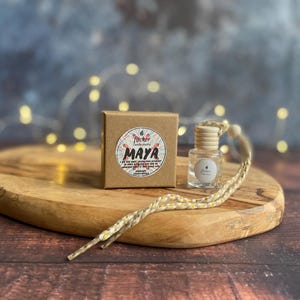 Könnte beinhalten: Eine kleine, klare Glasflasche mit einem Holzverschluss und einer dekorativen Schnur, neben einer kleinen braunen Schachtel. Die Schachtel hat ein rundes Etikett mit dem Wort "MAYA" und anderem Text. Die Artikel befinden sich auf einem Holzservierbrett.