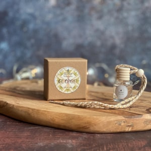 May include: A small, clear glass bottle with a wooden cap and a woven cord, next to a small brown box with a circular label that reads "Tucker Botanicals GEORGIE". The items are on a wooden serving board.