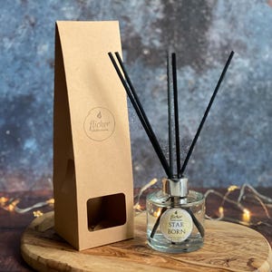 Puede incluir: Un difusor de aroma de vidrio transparente con cañas negras y una caja de cartón marrón. La botella tiene una tapa plateada y una etiqueta que dice "Flicker Candle Co. Star Born".