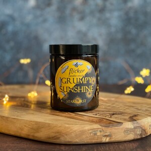 May include: A brown glass candle jar with a black lid. The label is yellow with black text that reads "flicker candle studio GRUMPY SUNSHINE STARBURST".