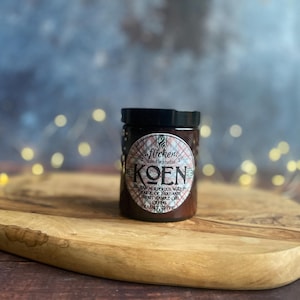 Może przedstawiać: Brązowy słoik ze świecą z czarną pokrywką stoi na drewnianej desce do krojenia. Słoik ma etykietę ze słowem "KOEN" i innym tekstem. Tło jest rozmyte ze światłami bokeh.