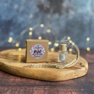 Könnte beinhalten: Eine kleine, klare Glasflasche mit Holzverschluss und geflochtener Kordel, neben einer kleinen braunen Schachtel. Die Schachtel hat ein farbenfrohes rundes Etikett mit dem Wort "RUE" und anderem Text. Die Artikel befinden sich auf einem Holzschneidebrett.