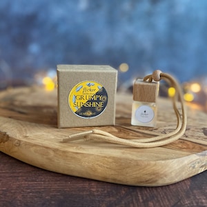 Może przedstawiać: Mała, przezroczysta butelka szklana z drewnianym korkiem i brązowym sznurkiem. Butelka jest oznaczona etykietą "flicker candle co. GRUMPY SUNSHINE STARDUST". Butelka stoi na drewnianej powierzchni.