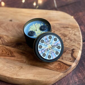 May include: A candle in a black tin with a blue wax interior, decorated with two large black buttons and small yellow sprinkles. The lid features the text "Coraline Strawberry & Chocolate Cake" and button illustrations.
