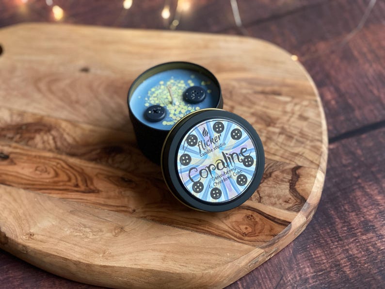 May include: A candle in a black tin with a blue wax interior, decorated with two large black buttons and small yellow sprinkles. The lid features the text "Coraline Strawberry & Chocolate Cake" and button illustrations.