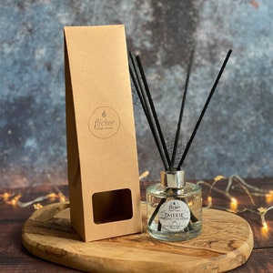 Könnte beinhalten: Ein Diffusor aus klarem Glas mit schwarzen Schilfrohren und einem braunen Karton. Die Flasche hat einen silbernen Verschluss und ein Etikett mit der Aufschrift "Emerie", "Natürlicher Schilfrohrdiffusor" und "Flicker Candle Studio".