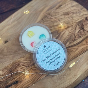 May include: A clear plastic container filled with white wax and topped with colorful house-shaped wax melts. The container has a label that reads "flicker candle studio" and "Your Island Name Here" with the text "Nook's Deserted Island Getaway Package Peach".