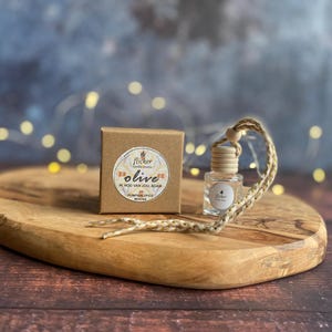 Könnte beinhalten: Eine kleine, klare Glasflasche mit einem Holzverschluss und einer geflochtenen Kordel, neben einer kleinen braunen Schachtel. Die Schachtel hat ein rundes Etikett mit dem Wort "Olive" und anderem Text. Die Artikel befinden sich auf einem Holzschneidebrett.