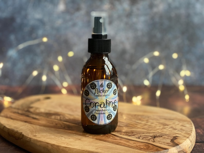 May include: A brown glass spray bottle with a black cap and a label that reads "Coraline Strawberry & Chocolate Cake" from Flicker Candle Studio. The bottle sits on a wooden cutting board. Fairy lights are in the background.