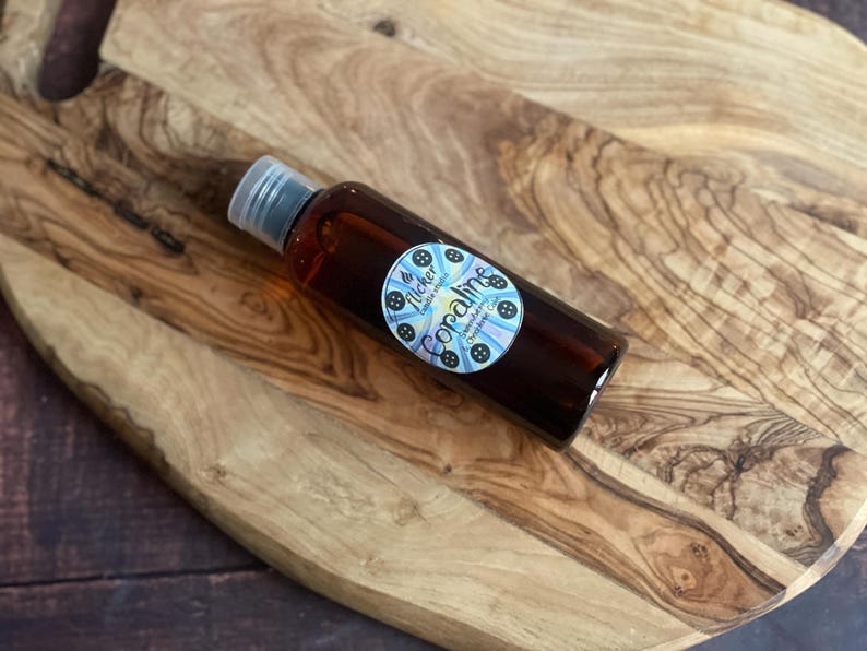 Pu&ograve; includere: Una bottiglia di vetro marrone con tappo argentato, riempita di liquido scuro, &egrave; appoggiata su un tagliere di legno. La bottiglia ha un'etichetta circolare con la scritta "Coraline" e un disegno blu e bianco.