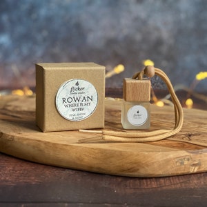 May include: A brown cardboard box with a white sticker that says "Flicker Candle Studio Rowan Where Is My Wife? Pine, Snow & Mint" and a small wooden car air freshener with a white sticker that says "Flicker Candle Studio" on a wooden surface.