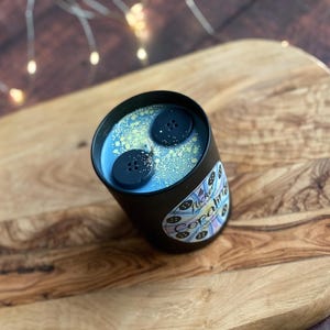 May include: A black candle tin with a light blue wax and two dark blue button-shaped wax melts. The candle has a label with the text "Tucker & Co. Opalite" and is set on a wooden cutting board.