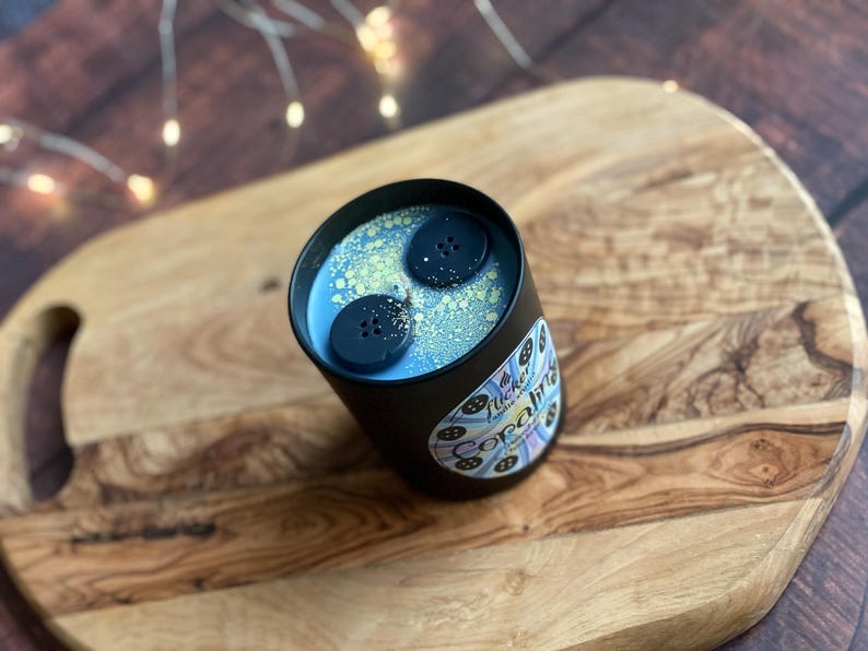 May include: A black candle tin with a light blue wax and two dark blue button-shaped wax melts. The candle has a label with the text "Tucker & Co. Opalite" and is set on a wooden cutting board.