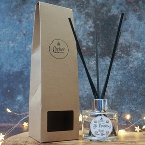 May include: A clear glass diffuser bottle with black reeds and a silver collar, next to a tall, brown cardboard box. The bottle has a label that reads "The Pumpkin King" and "flicker candle studio". The background is a textured blue surface.
