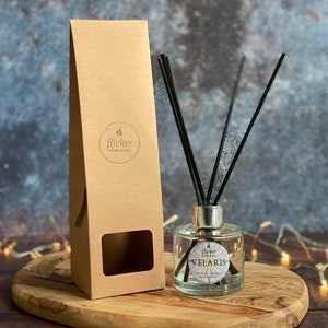 Könnte beinhalten: Ein Diffusor aus klarem Glas mit schwarzen Schilfrohren und einem silbernen Verschluss. Die Flasche trägt ein Etikett mit der Aufschrift "Flicker Candle Studio" und "Velaris" mit einem Sternmuster. Der Diffusor ist in einer braunen Pappbox mit einem Fenster verpackt.