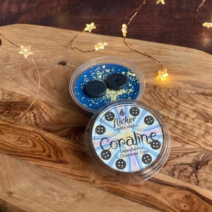 Könnte beinhalten: Zwei Wachsschmelzbehälter auf einem Holzbrett. Ein Behälter hat blaue Wachs mit zwei schwarzen Knöpfen. Der andere Behälter hat ein Etikett mit der Aufschrift "Coraline Strawberry & Chocolate Cake" mit Knöpfen. Lichterketten sind im Hintergrund.
