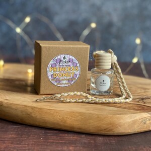 May include: A small, clear glass bottle with a wooden cap and a braided cord, next to a brown cardboard box. The box has a circular label with the words "Princess Donut" and "Flicker Candle Studio". The items are on a wooden tray.