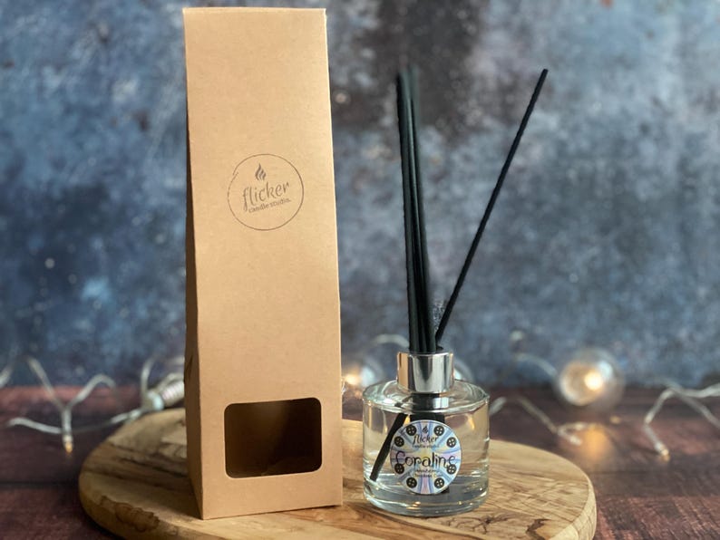 Pu&ograve; includere: Un diffusore in vetro trasparente con canne nere e un colletto argentato, accanto a una scatola di cartone marrone. La bottiglia ha la scritta "Coraline". La scatola ha il logo "flicker candle studio".