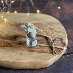 Può includere: Una piccola bottiglia di vetro trasparente con tappo in legno e cordino beige, probabilmente un diffusore di oli essenziali. Poggia su un tagliere di legno con venature naturali. Luci di fata sullo sfondo.