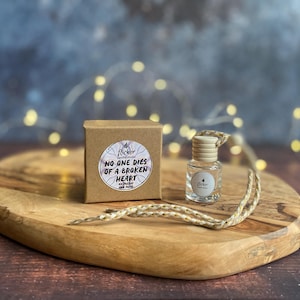 Puede incluir: Una pequeña botella de vidrio transparente con tapón de madera y cuerda de yute, junto a una pequeña caja marrón. La caja tiene una pegatina que dice "NO ONE DIES OF A BROKEN HEART". Sobre una tabla de madera.