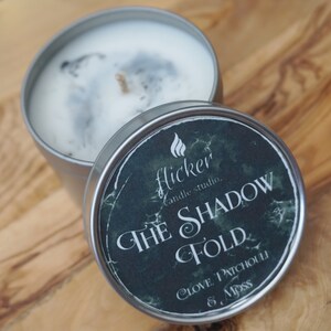 May include: A silver candle tin with a black and green label that reads "Flicker Candle Studio. The Shadow Fold. Clove, Patchouli & Moss".