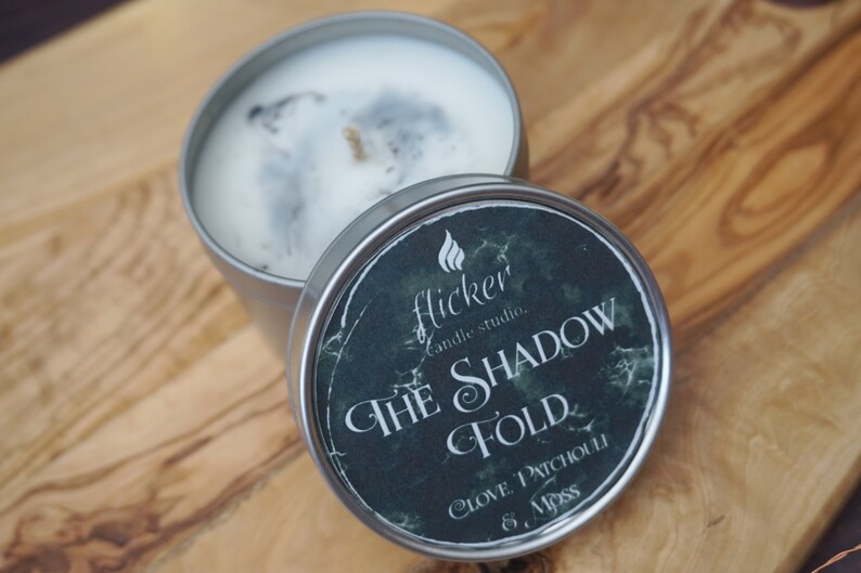 May include: A silver candle tin with a black and green label that reads "Flicker Candle Studio. The Shadow Fold. Clove, Patchouli & Moss".