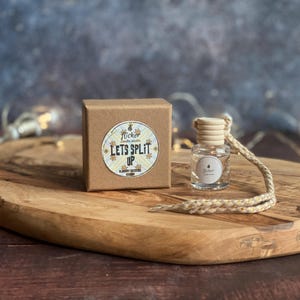 May include: A brown cardboard box and a small glass bottle with a wooden cap are displayed on a wooden oval board. The box has a sticker that reads "LET'S SPLIT UP". The bottle has a string attached.