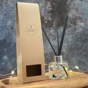 Puede incluir: Una botella de difusor de vidrio transparente con cañas negras y una etiqueta que dice "Warm Pumpkin Spice" y "Flicker Candle Studio". La botella está sobre una superficie de madera con una caja de papel marrón detrás.