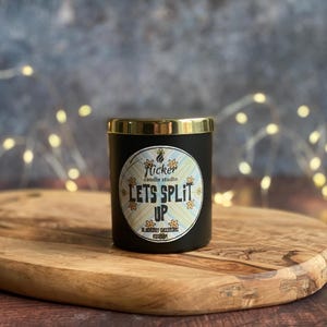 Könnte beinhalten: Eine schwarze Kerze mit goldenem Rand und dem Aufdruck "Lets Split Up" von Flicker Candle Studio. Die Kerze steht auf einem Holzbrett. Der Hintergrund ist eine verschwommene Mischung aus Farben und Lichterketten.