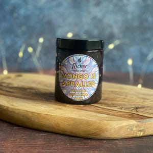 Op de afbeelding: Een donkere pot met een zwart deksel, met een kleurrijk etiket met de tekst "flicker candle studio" en "MONGO IS APPALLED" met "PEACH & PISTACHIO TART". De pot staat op een houten snijplank.