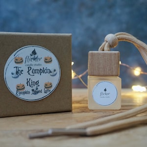 Könnte beinhalten: Eine kleine, quadratische Glasflasche mit einem Holzdeckel und einer beigefarbenen Kordel, neben einer braunen Schachtel mit einem runden Aufkleber mit der Aufschrift "The Pumpkin King Pumpkin Spiced Latte". Die Flasche hat ein weißes Etikett mit dem Wort "flicker".