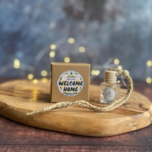 Könnte beinhalten: Ein Holzbrett zeigt eine kleine Pappschachtel und eine Glasflasche mit einem Holzdeckel. Auf der Schachtel befindet sich ein Aufkleber mit der Aufschrift "Welcome Home". An der Flasche ist eine geflochtene Kordel befestigt. Im Hintergrund befinden sich Lichterketten.