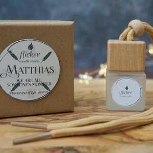 Könnte beinhalten: Ein brauner Karton mit einem weißen Aufkleber, auf dem "flicker candle studio" und "Matthias We are all someone's monster Peppermint & Eucalyptus" steht. Neben dem Karton befindet sich eine kleine, durchsichtige Glasflasche mit einem Holzdeckel und einer beigefarbenen Schnur. Die Flasche hat einen weißen Aufkleber mit der Aufschrift "flicker candle studio".