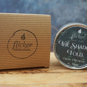May include: A small, round, metal candle tin with a black and green label. The label reads "flicker candle studio. THE SHADOW FOLD CLOVE. PATCHOULI 6. Moss".