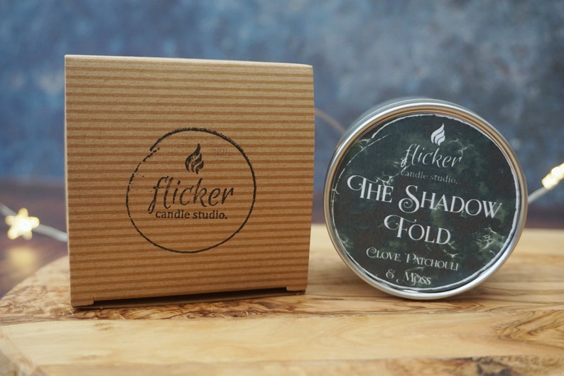 May include: A small, round, metal candle tin with a black and green label. The label reads "flicker candle studio. THE SHADOW FOLD CLOVE. PATCHOULI 6. Moss".