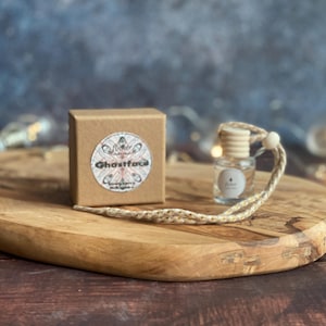 May include: A small glass bottle with a wooden cap and a woven cord, alongside a small, brown box. The box features a circular label with the text "Ghostface" and a decorative design. Both items are placed on an oval wooden tray.