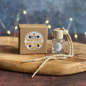 May include: A small, clear glass bottle with a wooden cap and a decorative string, next to a brown box. The box has a circular sticker with the text "YOU WILL NOT BREAK ME" and the words "amber, anise & mint". The items are on a wooden tray.