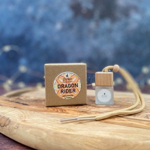 Może przedstawiać: Małe brązowe pudełko z napisem "Flicker Candles" i "Dragon Rider Cedarwood & Tonka". Obok pudełka znajduje się mała szklana butelka z drewnianym korkiem i beżowym sznurkiem. Butelka ma napis "Flicker".