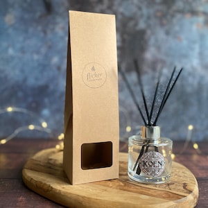 Könnte beinhalten: Eine klare Glas-Diffusorflasche mit schwarzen Schilfrohren und einem silbernen Kragen, beschriftet mit "KOEN", steht auf einem Holztablett neben einer hohen, braunen Pappschachtel mit einem Ausschnitt und dem Logo "flicker candle studio". Der Hintergrund ist verschwommen blau und braun.