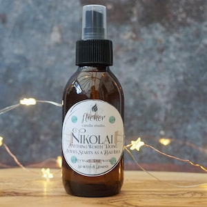 Puede incluir: Una botella de vidrio marrón con un pulverizador negro y una etiqueta blanca que dice "flicker candle studio NIKOLAI ANYTHING WORTH DOING ALWAYS STARTS AS A BAD IDEA" con un diseño verde y blanco. La botella está sobre una superficie de madera con una cadena de luces blancas con bombillas en forma de estrella.