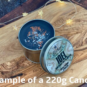 May include: A 220g candle in a silver tin with a black wax top. The candle is decorated with silver and orange glitter. The label on the tin reads "Flicker Candle Studio" and "JOEL DEAD PEOPLE CAN'T BE INFECTED COFFEE, CARAMEL & TOBACCO".