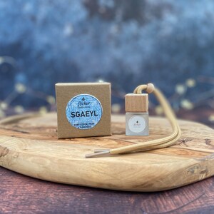 Może przedstawiać: Brązowe pudełko kartonowe z białą etykietą, na której widnieje napis "flicker candle studio" oraz "SGAEYL" czarnym kolorem. Etykieta zawiera również napis "sweet cocoa, pear & tobacco". Mała szklana butelka z drewnianym korkiem i brązowym sznurkiem leży na drewnianej powierzchni.