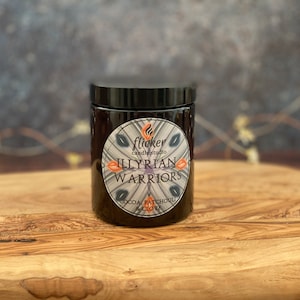 Puede incluir: Un tarro de vela de vidrio negro con tapa negra. La etiqueta dice "flicker candle studio Illyrian Warriors Cocoa Patchouli Clove".