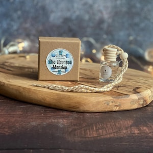 May include: A clear glass air freshener bottle with a wooden cap and a beige string, beside a small brown box with a blue and white label that reads "The Haunted Mansion". The items are on a wooden tray.